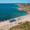 Spiaggia sabbiosa con ombrelloni blu e bianchi, acqua limpida e scogliere in lontananza.