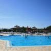 Persone che nuotano in una piscina con lettini, ombrelloni di paglia e cielo sereno.