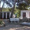 Un'area all'aperto con panchina bianca, due edifici semplici e alberi intorno.