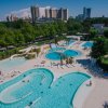 Complesso di piscine con scivoli e lettini, circondato da alberi e edifici sullo sfondo.