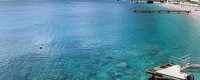 Mare cristallino con un molo in legno a destra e spiaggia in lontananza.