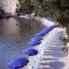 Spiaggia ciottolosa con ombrelloni blu allineati lungo la riva.