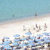 Spiaggia con lettini e ombrelloni blu disposti ordinatamente, persone che camminano lungo la riva.