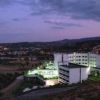 Complesso di edifici illuminato di notte con colline e città sullo sfondo.