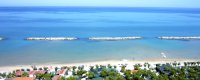 Spiaggia con mare limpido e barriere frangiflutti in lontananza.