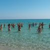 Persone fanno esercizi in mare con le braccia alzate sotto un cielo azzurro.