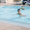 Un bambino in piscina con occhialini e braccioli, altri nuotatori sullo sfondo.