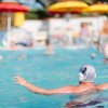 Un uomo in piscina gioca a pallanuoto, sfondo con ombrelloni colorati.