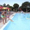 Papà lancia una bambina in piscina; ombrelloni colorati intorno.