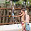 Due bambini giocano con arco e freccia giocattolo, vicino a una staccionata di legno.