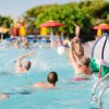 Persone che giocano a pallavolo in piscina con ombrelloni colorati sullo sfondo.