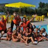 Gruppo di bambini e adulti sorridenti vicino a una piscina sotto ombrelloni gialli.