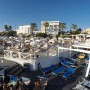 Spiaggia affollata con lettini blu, ombrelloni bianchi, e edifici bianchi sullo sfondo.