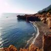 Spiaggia di ciottoli con scogliere rosse, mare blu e sole che riflette sull'acqua.